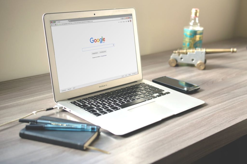 macbook-air-on-grey-wooden-table-67112 A neat workspace featuring a laptop displaying Google search, a smartphone, and a notebook on a wooden desk.